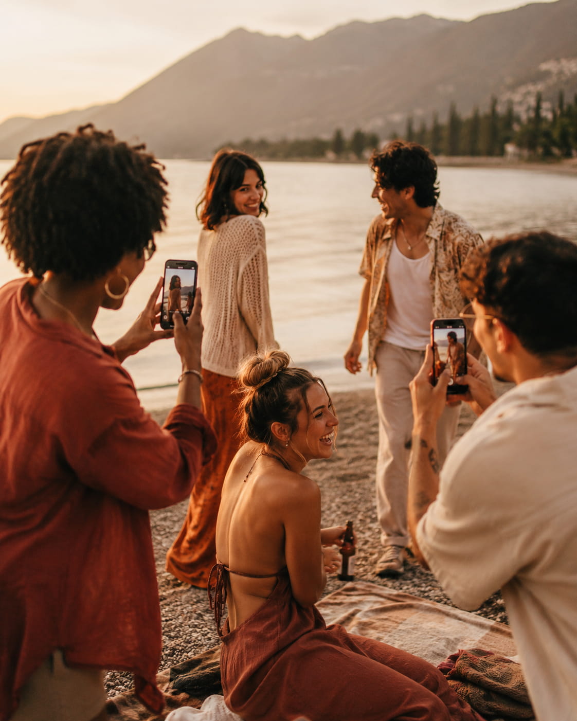 Group of friends laughing at golden hour by a mountain lake, some holding phones and cameras