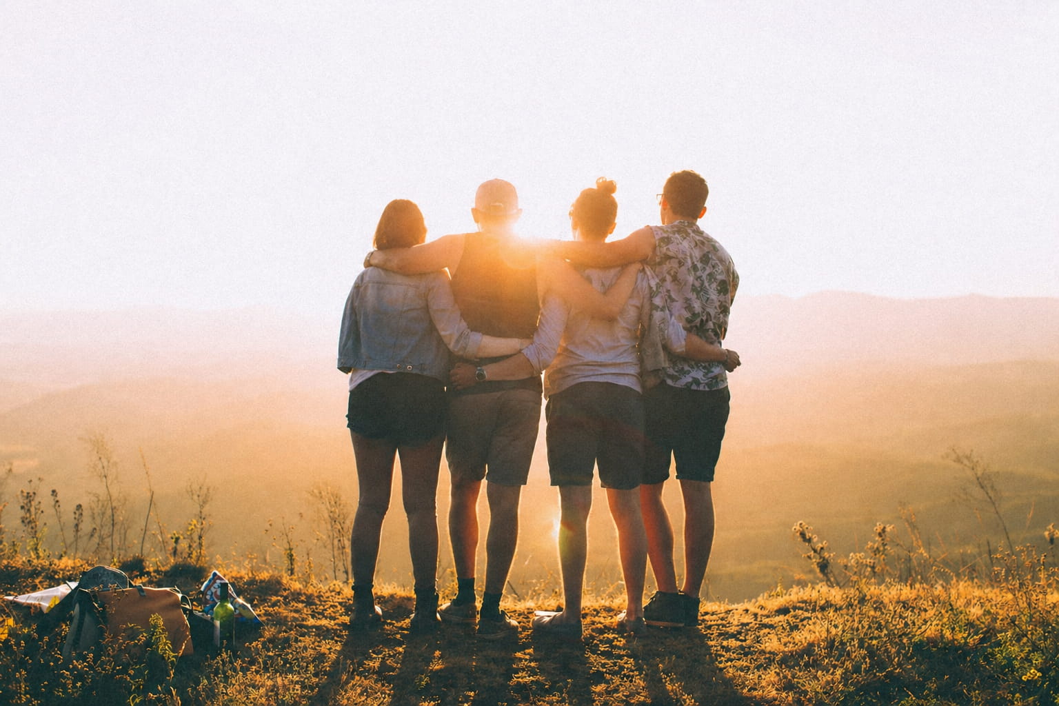 Group of friends with arms around each other watching a golden sunset from behind